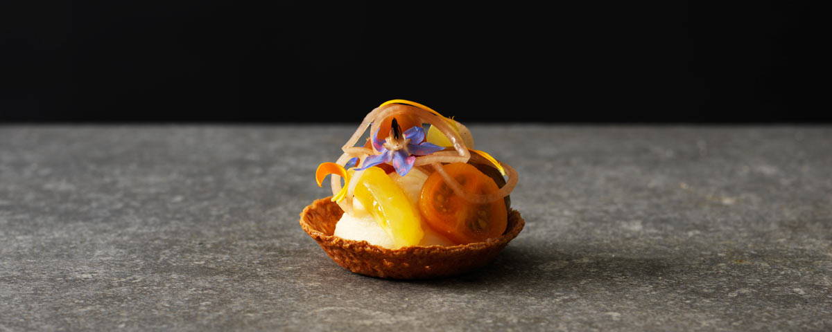 Billede af en lille sprød tartelet, med fyld af Gammel Knas Creme og toppet med tomat. 